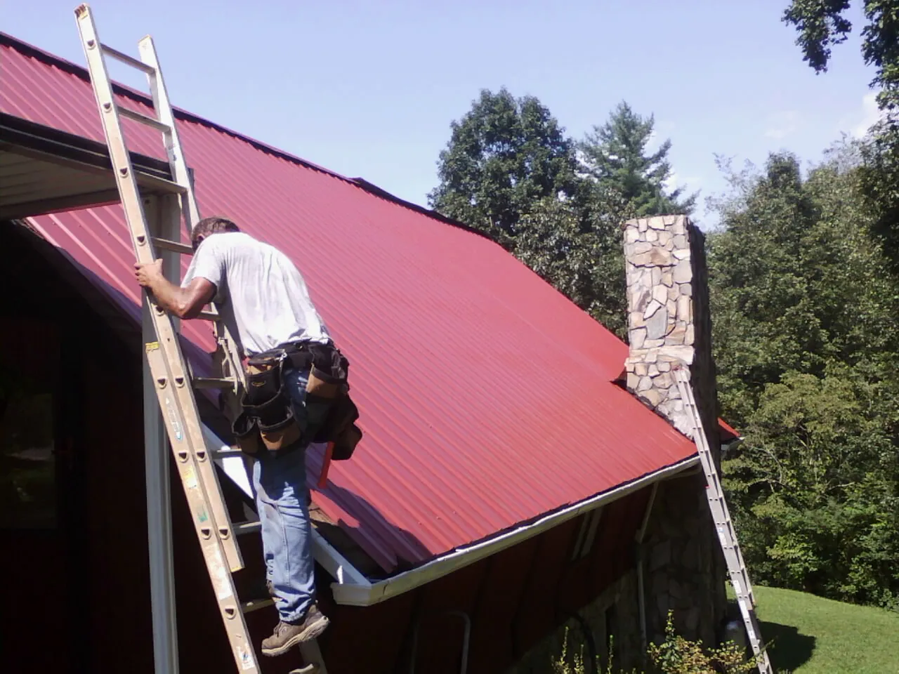 Roofer on a ladder beside a red metal roof and stone chimney — Soffit & Fascia Repair by Roofing Brothers in North Wilkesboro